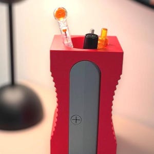 Pode incluir: Um porta-lápis vermelho em forma de apontador de lápis com uma lâmina de afiar cinza. O porta-lápis tem um pequeno sinal de mais na frente e contém alguns lápis.