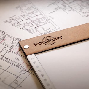 May include: A brown leather drafting tool with the text "RotoRuler" on the top. The tool is lying on a white sheet of paper with architectural drawings.
