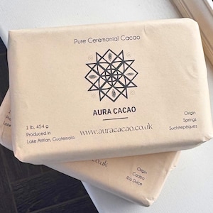 以下が含まれることがあります： ベージュ色の紙に包まれた、Aura Cacaoの長方形のパッケージが2つ。パッケージは積み重ねられており、上部には「Pure Ceremonial Cacao」の文字と幾何学模様が表示されています。「AURA CACAO」の文字と重量454gも表示されています。