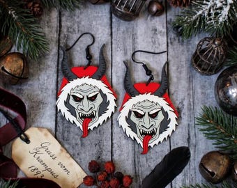 May include: Two Krampus earrings with a red, white, and grey Krampus face, horns, and a red tongue. The earrings have black hooks. The image includes a tag with text and festive decorations, including fir branches.
