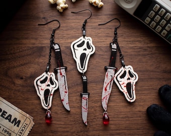 May include: Black and white Scream movie-themed earrings. Each earring features a Ghostface mask and a knife with red blood splatter. A red teardrop bead hangs from each earring. The earrings are on a wooden surface.