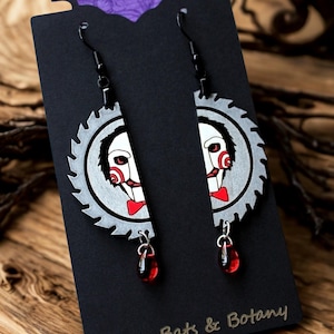 May include: A pair of unique earrings featuring a saw blade design with a white and red face graphic. The earrings have a red teardrop charm and are displayed on a black card with the text "Bones Bats & Botany".