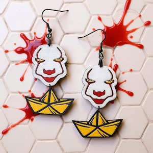 Op de afbeelding: Een paar Halloween-oorbellen met een wit clowngezicht met rode accenten en een gele papieren boot. De oorbellen hangen aan zwarte haken, tegen een rode spetterachtergrond.