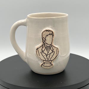 May include: A cream-colored ceramic mug with a smooth, glossy finish and a curved handle. Centered on the front is a raised, textured relief of a person in a formal suit and cravat, depicted from the chest up in a classic bust style.