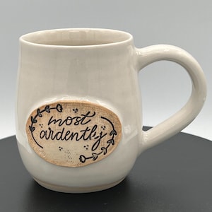 May include: A glossy off-white ceramic mug featuring a centered oval medallion with a tan, textured surface. The medallion displays the black handwritten text "most ardently" surrounded by a delicate black vine and leaf illustration.