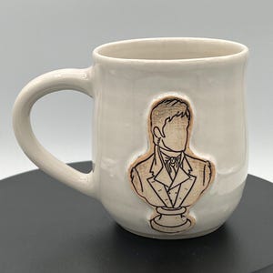 May include: A white ceramic mug with a glossy finish and a curved handle. The front features a raised, tan-colored relief of a person in a formal suit, depicted as a bust with a black outline. The figure has a collared jacket and a necktie.