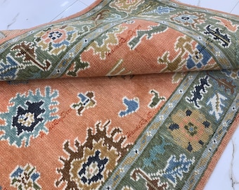 May include: A rolled-up area rug with a detailed, traditional design. The rug features a peach-coloured central field with repeating patterns in shades of blue, green, and brown. The border is a darker green with similar motifs.