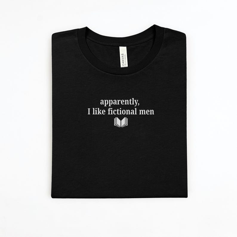 May include: Black t-shirt with the text "apparently, I like fictional men" in white, along with a small open book graphic. The shirt is folded neatly, showcasing the design.