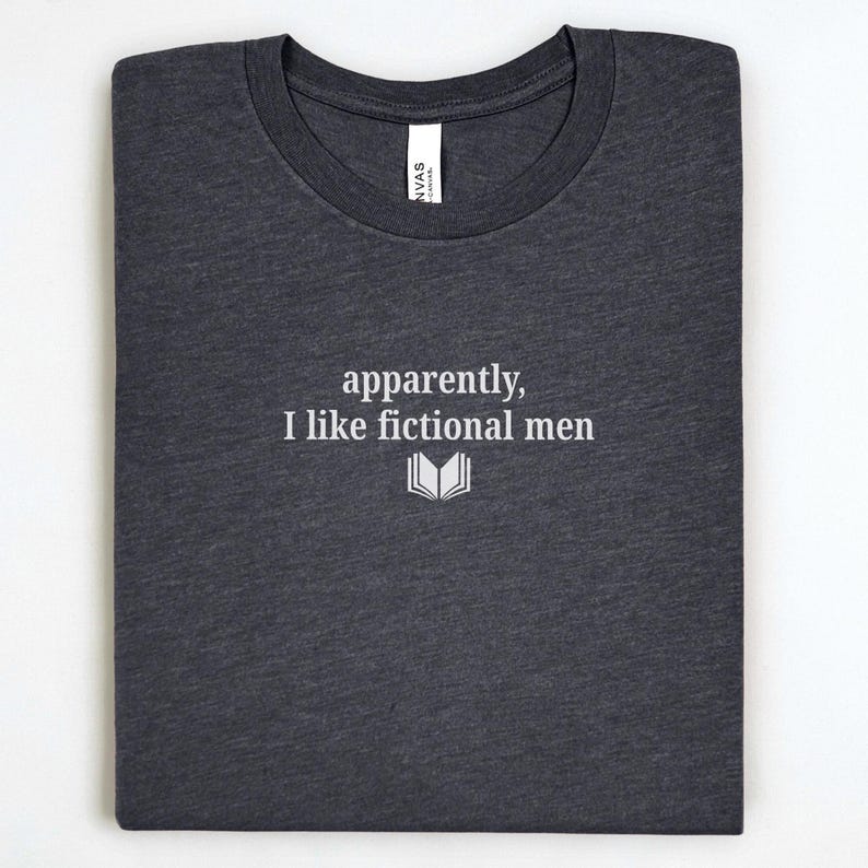 May include: Dark gray t-shirt with the text "apparently, I like fictional men" in white, along with a small open book graphic. The shirt is folded neatly, showcasing the design.