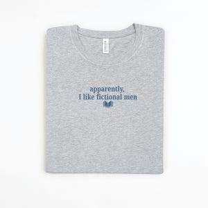 May include: A light gray t-shirt with the text "apparently, I like fictional men" in navy blue, along with a small book graphic. The shirt is folded neatly, with a white tag visible near the neckline.