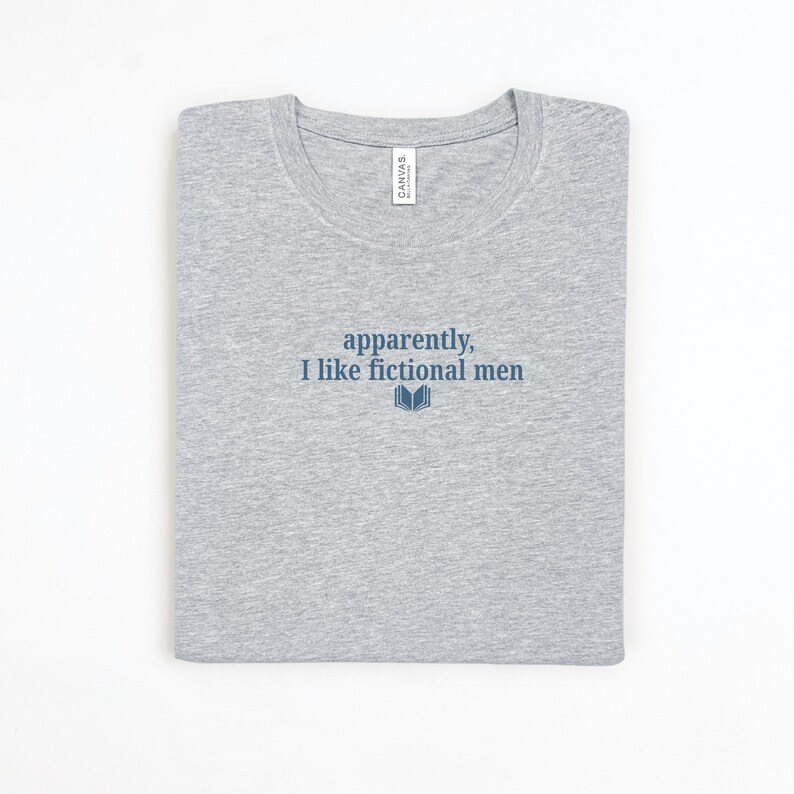 May include: A light gray t-shirt with the text "apparently, I like fictional men" in navy blue, along with a small book graphic. The shirt is folded neatly, with a white tag visible near the neckline.