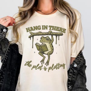 May include: Cream-colored t-shirt featuring a cartoon frog reading a book while hanging from a branch. The text "HANG IN THERE" is above the frog, and "the plot is plotting" is below. The frog is green with brown spots.