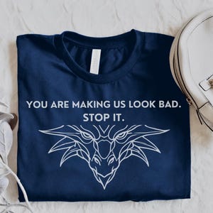 May include: A navy blue t-shirt with a white dragon graphic and the text "You are making us look bad. Stop it."