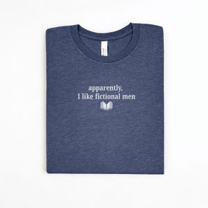 May include: A navy blue t-shirt with the text "apparently, I like fictional men" in white, along with a small open book graphic. The shirt is folded neatly on a white surface.