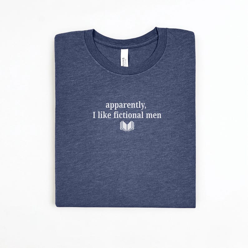 May include: A navy blue t-shirt with the text "apparently, I like fictional men" in white, along with a small open book graphic. The shirt is folded neatly on a white surface.