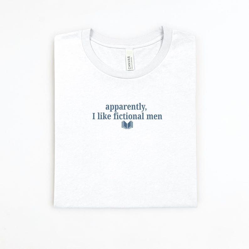 May include: White t-shirt with a crew neck and the text "apparently, I like fictional men" in blue, along with a small open book graphic. The shirt is folded neatly.