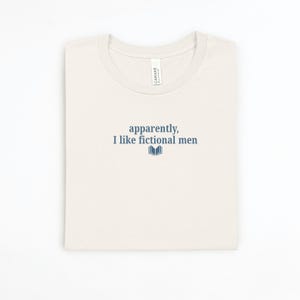 May include: Cream-colored t-shirt with the text "apparently, I like fictional men" in navy blue, along with a small book graphic. The shirt is folded neatly on a white surface.