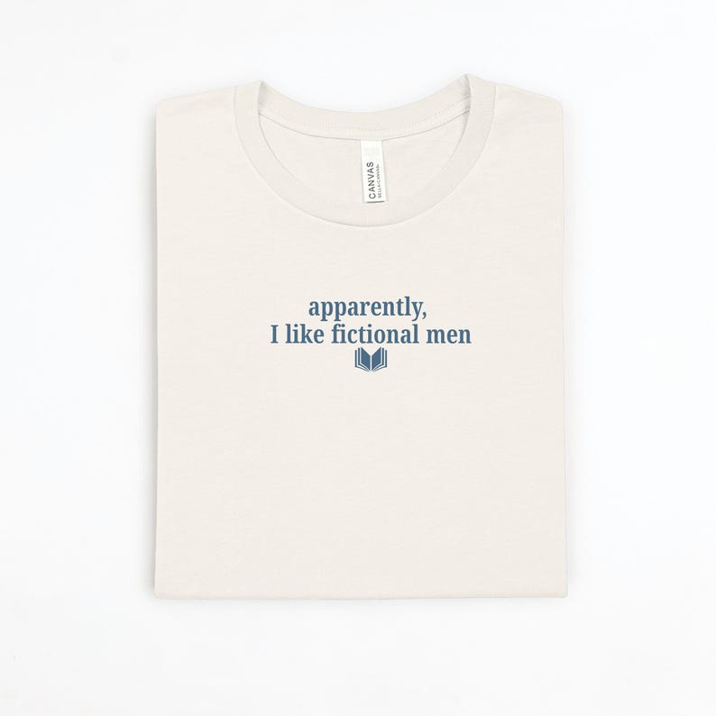 May include: Cream-colored t-shirt with the text "apparently, I like fictional men" in navy blue, along with a small book graphic. The shirt is folded neatly on a white surface.