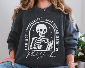 May include: Dark gray sweatshirt with a white graphic of a skeleton reading a book. The text reads "I am not dissociating, just vibing elsewhere" and "Plot Junkie". The sweatshirt is a casual style.