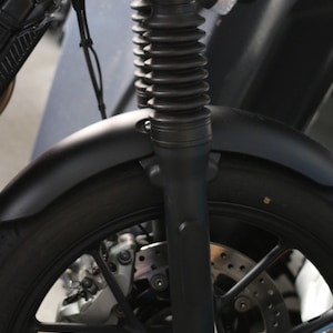 May include: A matte black motorcycle front end featuring a telescopic fork with black rubber gaiters, a curved front fender, a multi-spoke alloy wheel, and a ventilated disc brake system with a silver caliper mounted near the wheel hub.