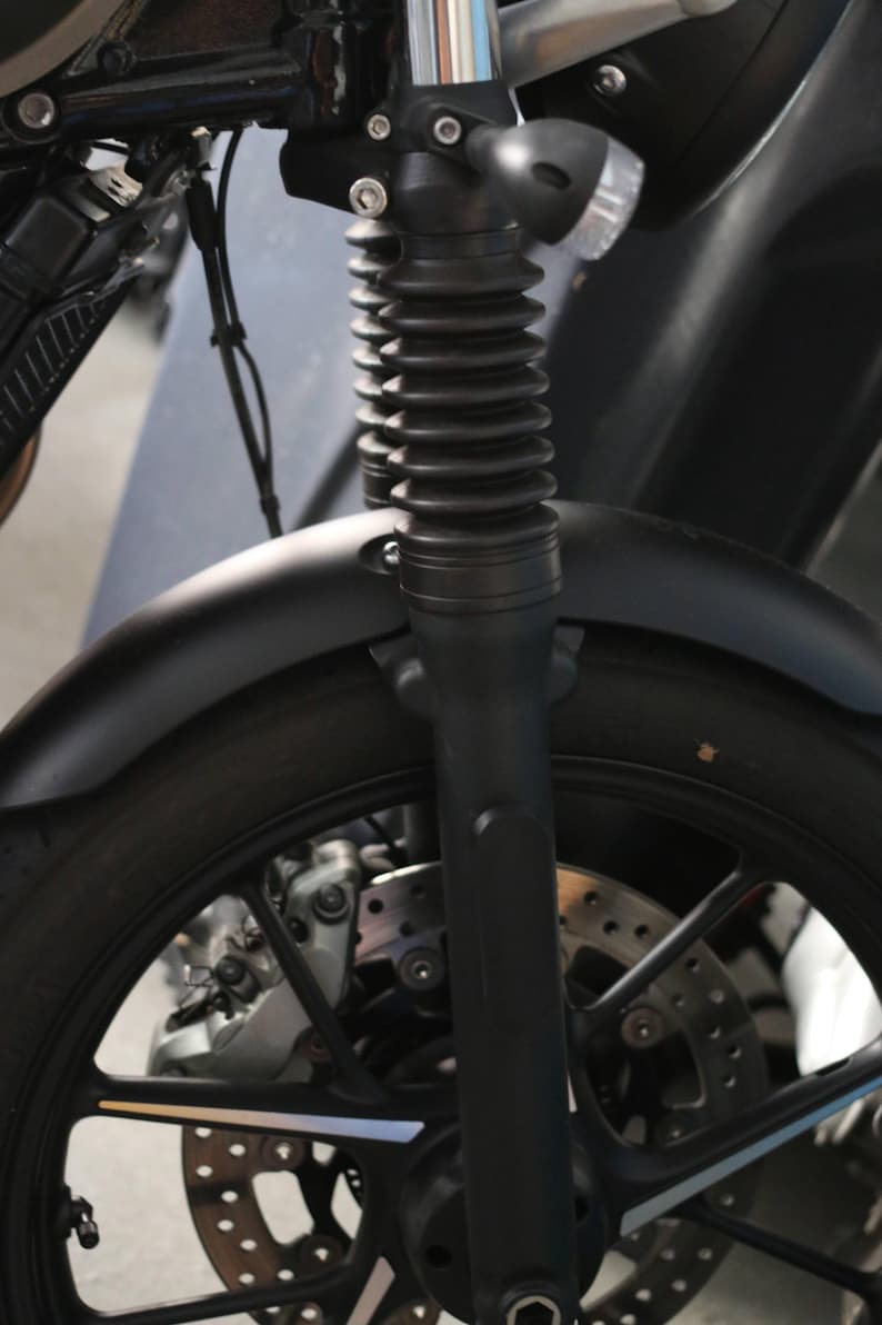 May include: A matte black motorcycle front end featuring a telescopic fork with black rubber gaiters, a curved front fender, a multi-spoke alloy wheel, and a ventilated disc brake system with a silver caliper mounted near the wheel hub.