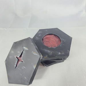 May include: A set of gray, coffin-shaped coasters with a distressed finish. One coaster has a star-shaped cutout revealing a red interior. The coasters are stacked, with one partially open, revealing a red, textured surface.