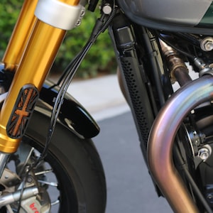 May include: A motorcycle front end featuring gold-colored forks, a black fender, and a black radiator guard with a Union Jack flag cutout design. An iridescent, heat-treated exhaust pipe curves near the engine block, with visible brake components.