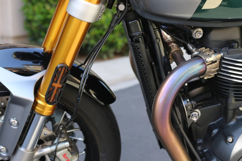 May include: A motorcycle front end featuring gold-colored forks, a black fender, and a black radiator guard with a Union Jack flag cutout design. An iridescent, heat-treated exhaust pipe curves near the engine block, with visible brake components.