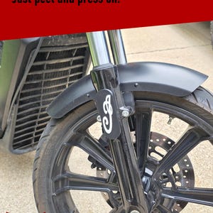 May include: A black motorcycle fork decal featuring a white stylized script logo applied to the front suspension. The text at the top reads Easy to apply! - Just peel and press on! The decal is positioned on the black fork of a motorcycle wheel.