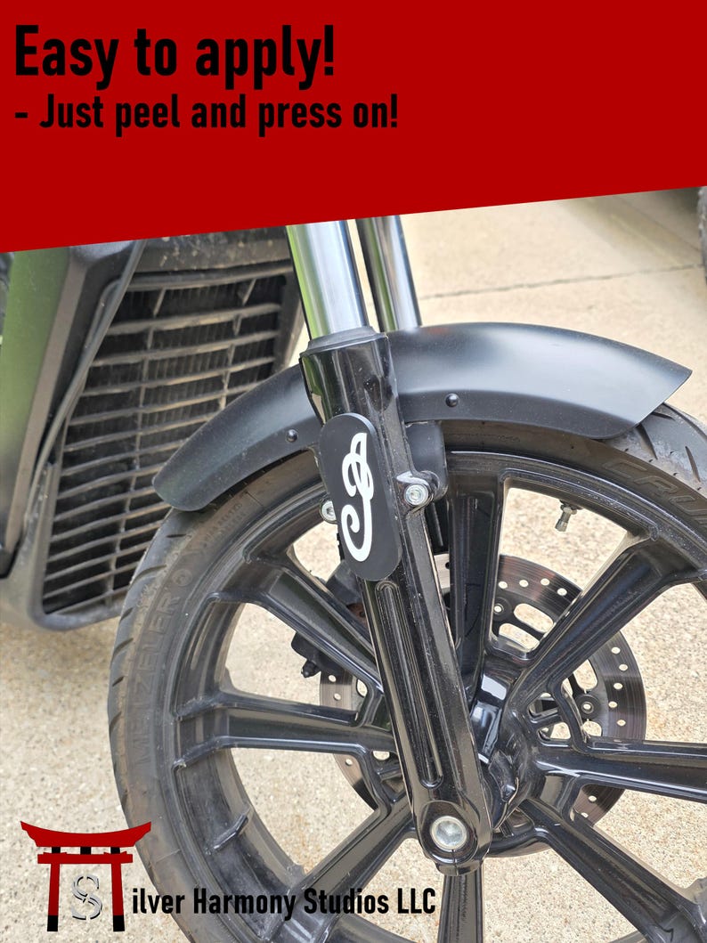 May include: A black motorcycle fork decal featuring a white stylized script logo applied to the front suspension. The text at the top reads Easy to apply! - Just peel and press on! The decal is positioned on the black fork of a motorcycle wheel.