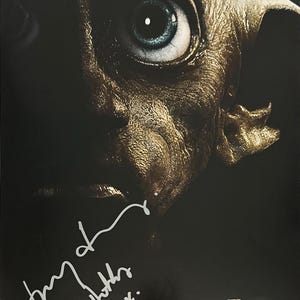 Pode incluir: Um cartaz com um close-up de Dobby de Harry Potter e as Relíquias da Morte Parte 1. A imagem mostra o rosto de Dobby com um olho grande e uma assinatura. O cartaz tem o título do filme e a frase "Nowhere Is Safe".