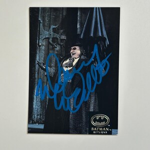 Może przedstawiać: Karta kolekcjonerska Batman Returns z postacią w kostiumie i niebieskim autografem. Karta ma ciemne tło z logo Batman Returns w prawym dolnym rogu. Postać uśmiecha się i ma na sobie czarny płaszcz.