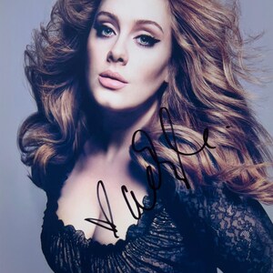 May include: A portrait of a woman with long, wavy auburn hair and striking blue eyes. She wears dark eye makeup and a black lace top. The image includes a signature in black ink.