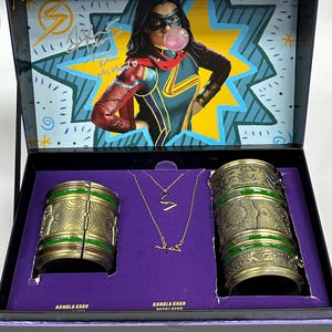 May include: A purple box containing a gold and green bracelet set. The box features a picture of Kamala Khan, also known as Ms. Marvel, in her superhero costume. The box also includes the text "Kamala Khan Ms. Marvel".