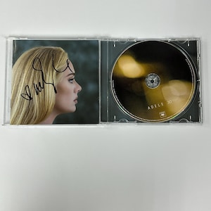 Puede incluir: Una caja de CD firmada con una foto de Adele en la portada y el título "Adele 30" en el CD.