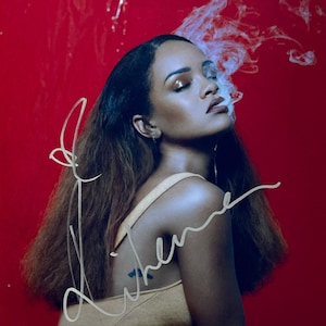 May include: A woman with long brown hair and a yellow dress exhales smoke against a red background. The image has a signature in white cursive script. A small blue tattoo is visible on the woman's back.