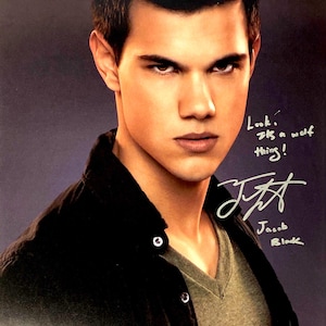 Puede incluir: Una fotografía en color de un joven con el pelo corto y castaño, que lleva una camisa verde oscuro y una chaqueta negra. Está mirando directamente a la cámara con una expresión seria. La imagen está firmada "Jacob Black" en tinta plateada. El texto "Look, it's a wolf thing!" está escrito en tinta negra.