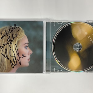 Puede incluir: Un CD firmado del álbum "25" de Adele con una foto de Adele en la portada. El CD está en una caja de plástico transparente.