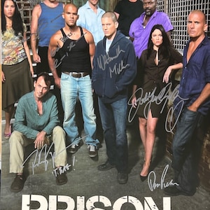 May include: A signed poster of the cast of the TV show Prison Break. The cast members are standing in a prison setting and are all wearing casual clothing. The poster is signed by the actors, including Wentworth Miller, Dominic Purcell, and Sarah Wayne Callies.