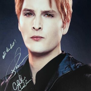 Puede incluir: Una fotografía firmada de un actor con el pelo rojo, que lleva una chaqueta negra con forro azul. El actor está mirando directamente a la cámara. La firma dice "All the best!" y "Cullen."