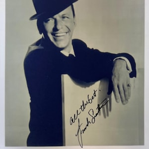 Peut inclure: Photographie en noir et blanc de Frank Sinatra portant un fedora et un costume, souriant. L'image comprend sa signature et les mots "All the best." Le texte "FRANK SINATRA" est en bas.