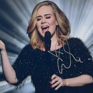 Peut inclure: Photographie d'une femme chantant dans un microphone sur scène. Elle porte un haut noir scintillant et a les cheveux blonds. L'image comprend une signature blanche.