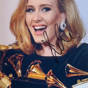 Puede incluir: Una mujer sonriente con cabello rubio y un top negro sostiene varios premios Grammy de oro. Lleva un pendiente de diamantes y tiene una firma en la imagen. Los premios son dorados y negros.
