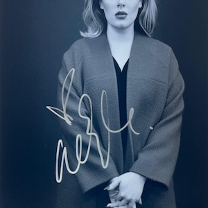 Puede incluir: Retrato de una persona con cabello rubio, vistiendo un abrigo gris sobre una blusa negra. La imagen tiene una firma en blanco. El fondo es de color azul oscuro.