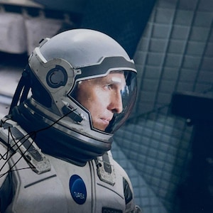 May include: A signed photograph featuring a person wearing a space suit and helmet. The suit has a NASA logo and the word "Interstellar" is visible. The background is a dark, futuristic interior.