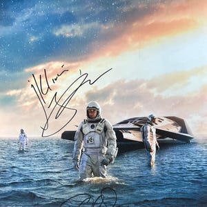 May include: A black and white movie poster featuring three astronauts in white spacesuits standing in a body of water. The astronauts are standing in front of a white spaceship. The sky is a dark blue with white stars and a light orange sunset. The poster is signed by two of the astronauts.