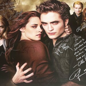 Puede incluir: Un grupo de actores de la saga de películas Twilight, incluyendo a Robert Pattinson, Kristen Stewart y Taylor Lautner, aparecen con sus autógrafos en un póster. El póster es una imagen en blanco y negro de los actores con un fondo oscuro.