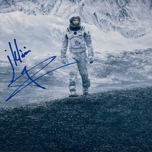 May include: A signed photograph featuring a person in a spacesuit standing on a rocky surface, with a snowy mountain backdrop. The signature is in blue ink. The image has a cool, blue-toned color palette.