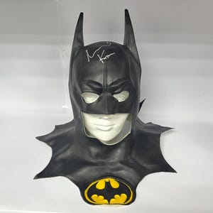 Puede incluir: Una máscara de Batman negra con un símbolo de murciélago amarillo. La máscara está firmada "M. Keaton".