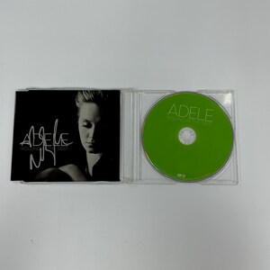 Può includere: Una custodia per CD con una foto in bianco e nero di Adele sulla copertina anteriore. La custodia per CD è aperta e il CD è visibile. Il CD è verde e ha il testo "Adele Rolling in the Deep" stampato su di esso.